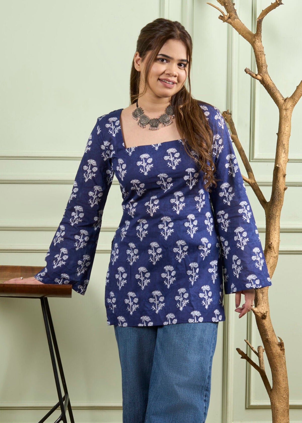 A woman wearing a navy blue long-sleeve kurta with a white floral block print pattern, paired with blue jeans, standing in front of a light green paneled wall next to a decorative tree branch and a wooden stool.