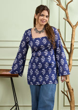 A woman wearing a navy blue long-sleeve kurta with a white floral block print pattern, paired with blue jeans, standing in front of a light green paneled wall next to a decorative tree branch and a wooden stool.