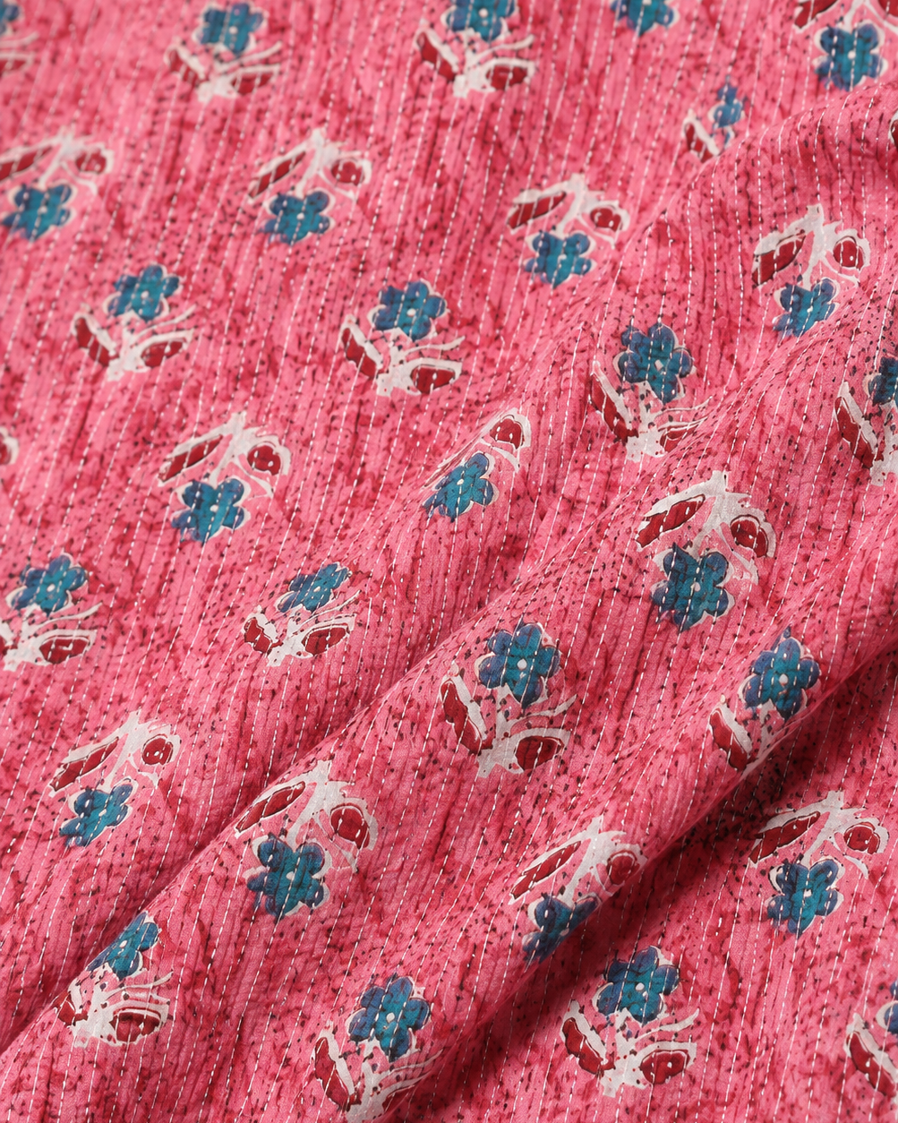 A close-up of a pink textured fabric with a repeating block-print pattern of small blue flowers and white leafy stems.