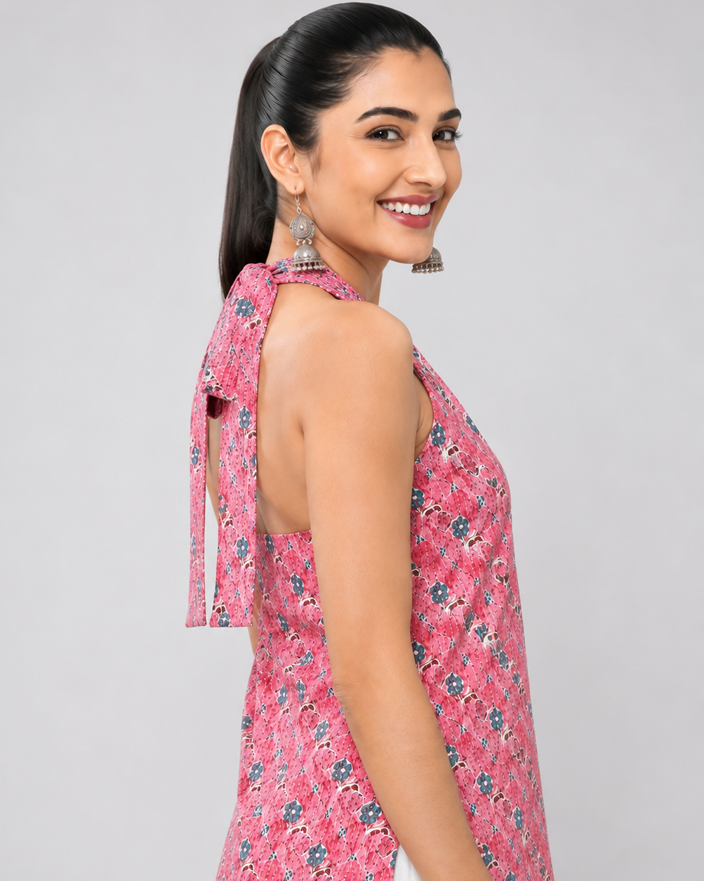 A woman modeling a sleeveless pink floral printed top with a tie-neck detail, shown from a side-back angle against a light gray studio background.