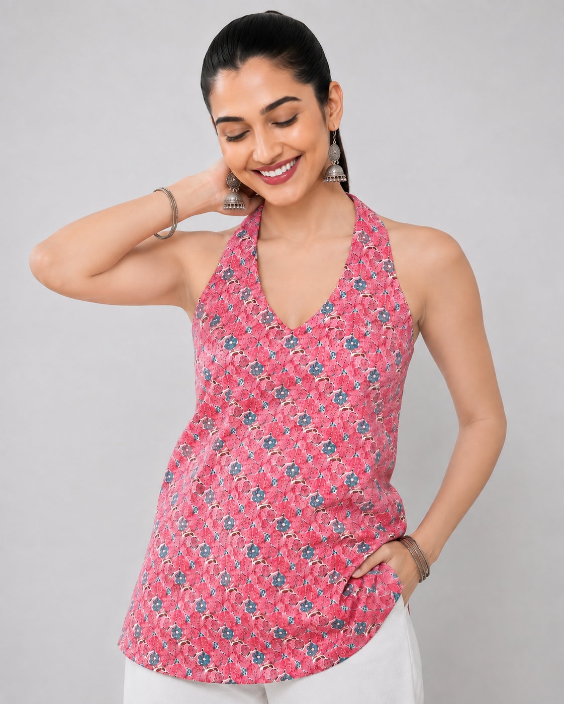 A woman modeling a pink sleeveless halter top with a blue and white floral pattern, paired with white pants, against a solid gray studio background.