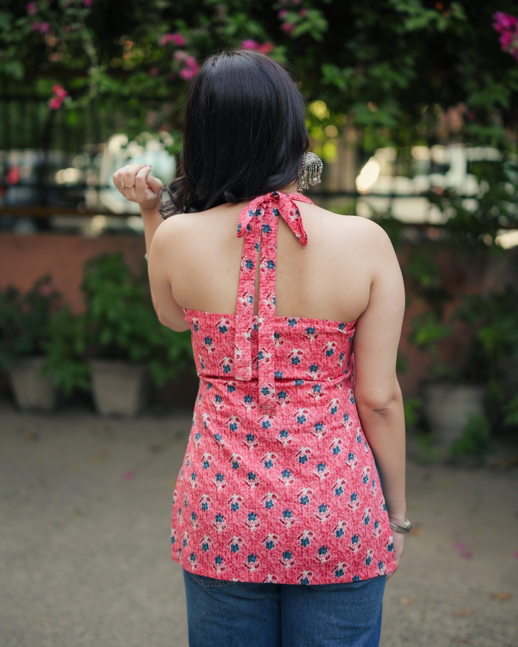A woman viewed from behind wearing a pink floral halter top and blue jeans, standing outdoors in front of green foliage and flowers.