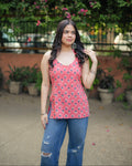 A woman wearing a red sleeveless tank top with a blue and black geometric pattern, paired with distressed blue jeans, standing outdoors in front of a green garden background.