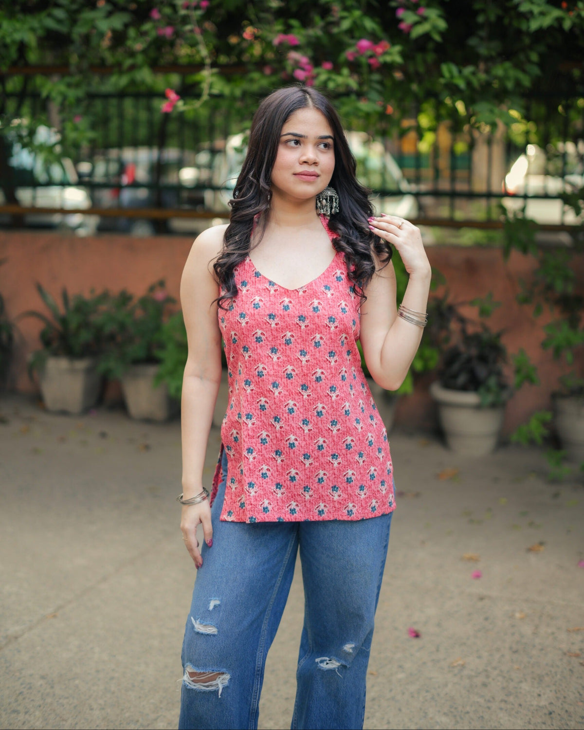 A woman wearing a red sleeveless tank top with a blue and black geometric pattern, paired with distressed blue jeans, standing outdoors in front of a green garden background.