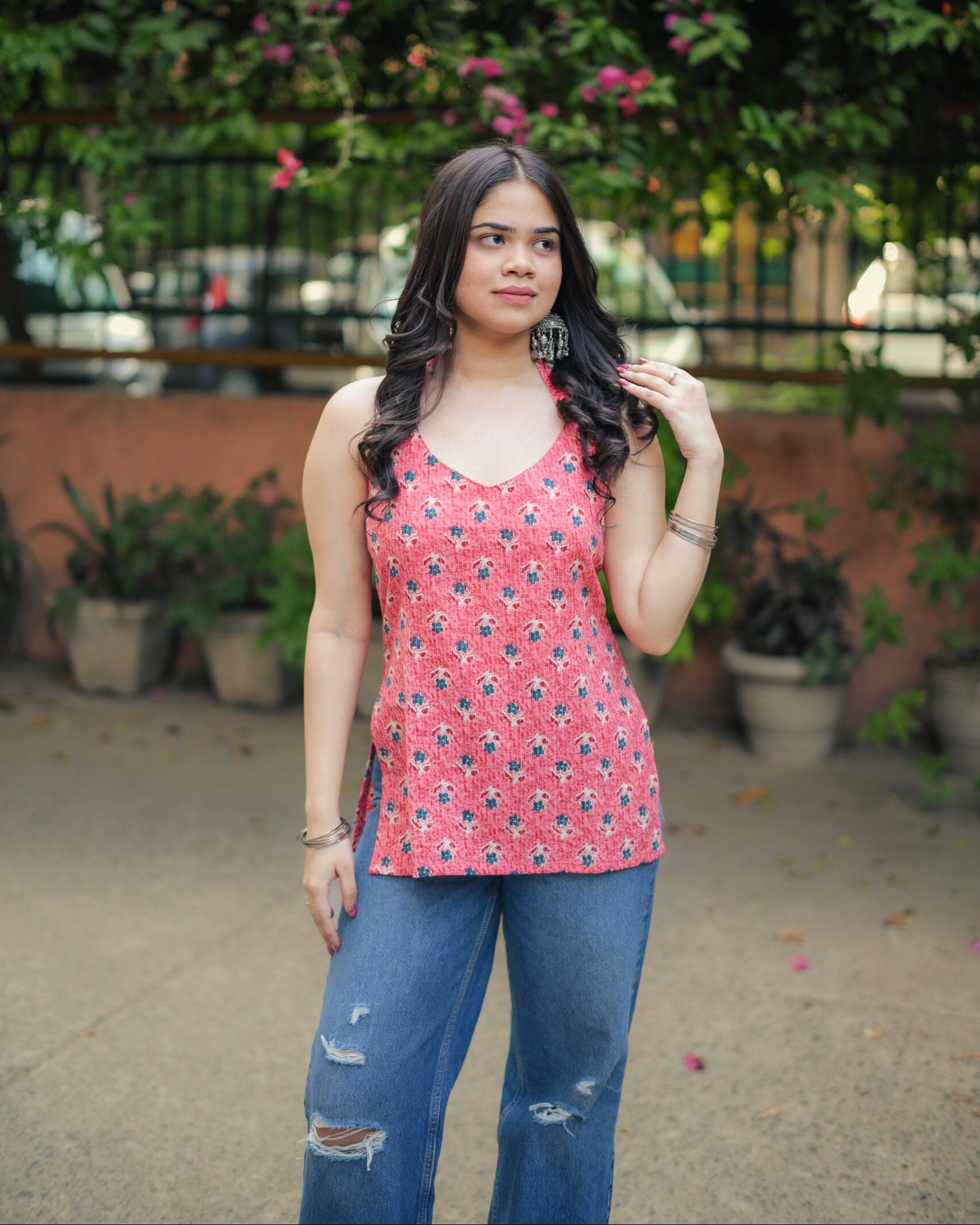 A woman wearing a red sleeveless tank top with a blue and black geometric pattern, paired with distressed blue jeans, standing outdoors in front of a green garden background.
